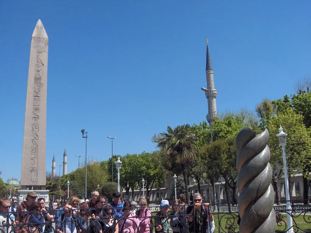 Önde Yılanlı Sütun'un tunç gövdesi, arkada Dikilitaş ve bir minare, parmaklık çevresinde toplanmış ziyaretçiler ve ağaçlar.