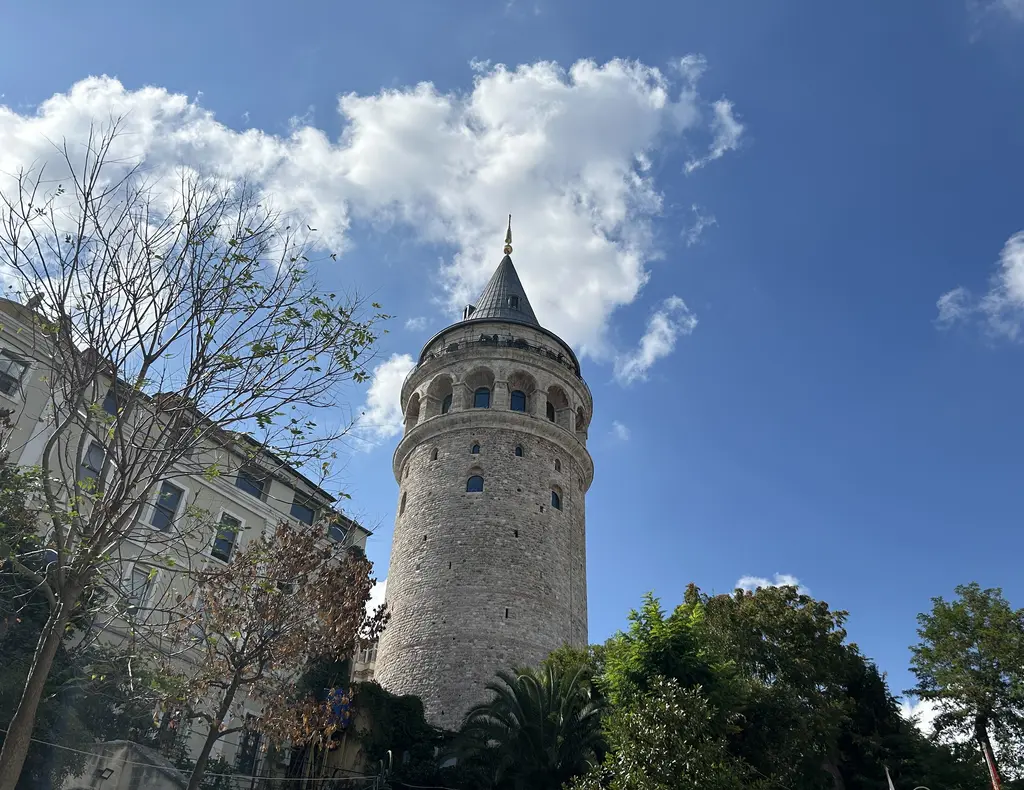 Galata Kulesi; konik külahı ile kemerli pencere sırası, gri taş gövdesi, önde yeşil ağaçlar ve arkada beyaz bulutlu mavi gökyüzü.