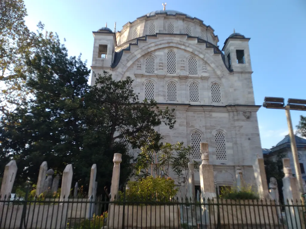 Ayazma Camii'nin haziresi; parmaklık ardında sıralanan mezar taşları, ağaçlar ve arkada caminin cephesi.