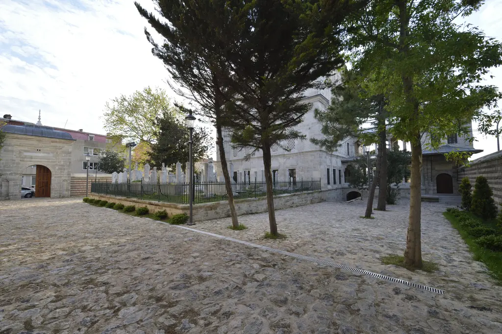 Ayazma Camii'nin avlusu; taş döşeli zemin, sıra ağaçlar, demir parmaklıklı yeşil alan ve uzakta avlu kapısı.