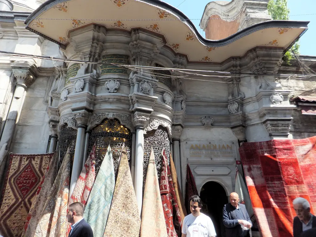 Nuruosmaniye Külliyesi'nin sebili; dalgalı oyma mermer saçak, tunç şebekeli yuvarlak pencereler, ince sütunlar ve önünde asılı duran halılar.
