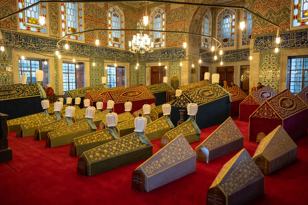 Sultan Ahmed Türbesi'nin içi; kırmızı halı üstünde sıralanan yaldız işlemeli sandukalar, çini kaplı duvarlar, renkli camlı pencereler ve asılı kandiller.