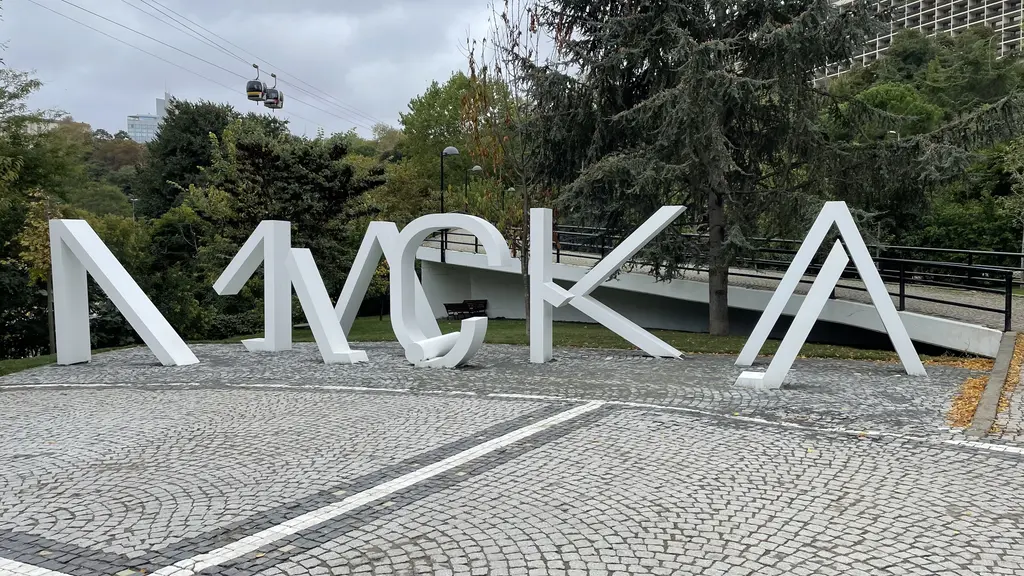 Maçka Demokrasi Parkı'nın girişinde taş döşeli meydanda dev beyaz "MAÇKA" harfleri, arkada ağaçlar ve gökyüzünde teleferik kabini.