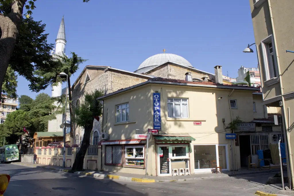 Kuzguncuk'ta sahil yolu köşesi; krem renkli köşe binası, dükkânlar, arkada kubbe ve minare, önde ağaçlar.