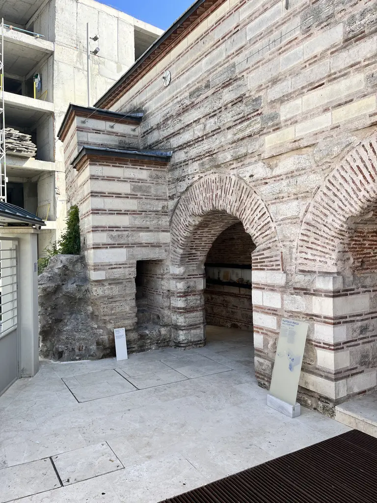 Çinili Hamam'da Kuyulu Avlu; almaşık taş ve tuğla duvarda iki büyük tuğla kemer, beyaz mermer zemin, bilgi panoları ve arkada bir bina.