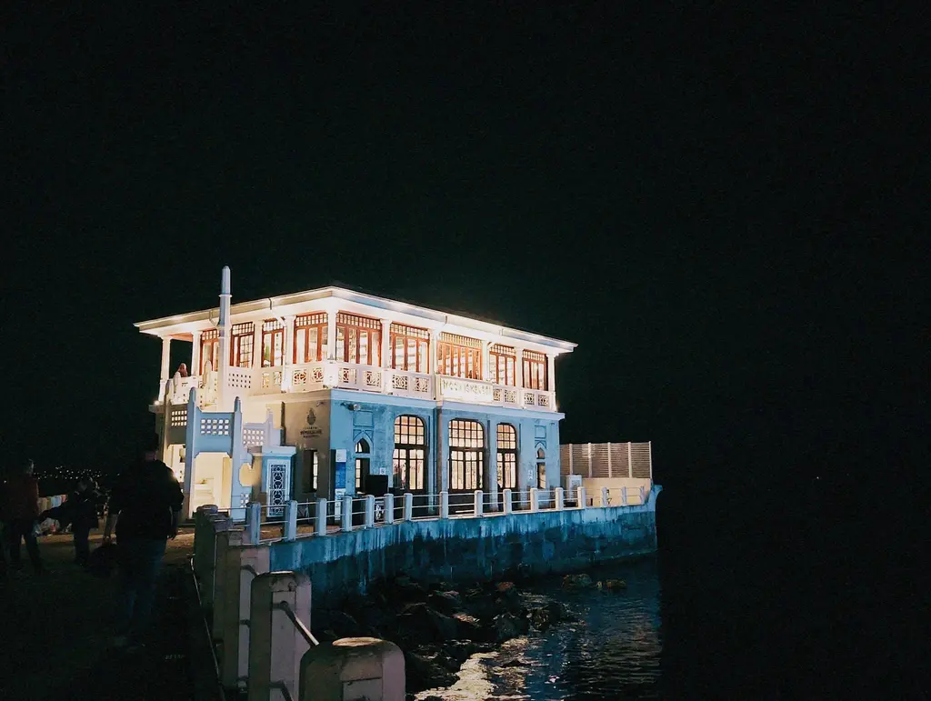 Gece Moda İskelesi; aydınlatılmış bina, ışıklı pencereler, mavi aydınlatılmış alt kat ve karanlık gökyüzü.