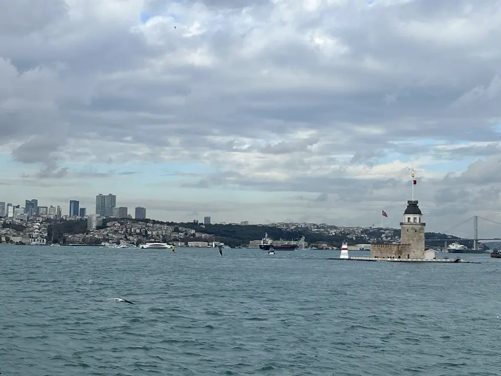 Kız Kulesi ve Avrupa yakası; bulutlu gökyüzü altında Boğaz, sağda küçük kule, arkada Beşiktaş ve Levent'in binaları.