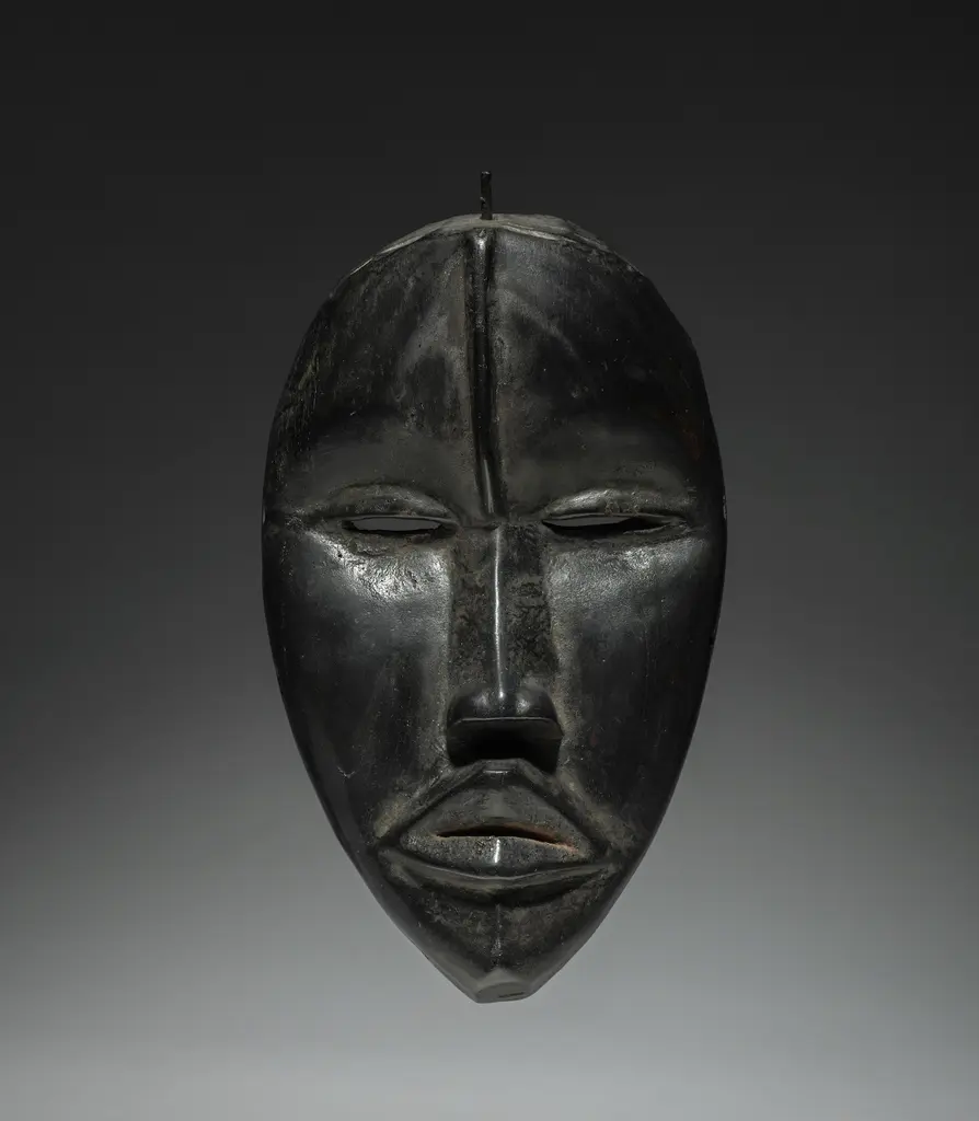 Koyu ahşaptan oyulmuş, parlatılmış geleneksel bir Batı Afrika maskesi (Dan tipi); ince uzun yüz, badem gözler ve dolgun dudaklar, gri stüdyo zemininde.