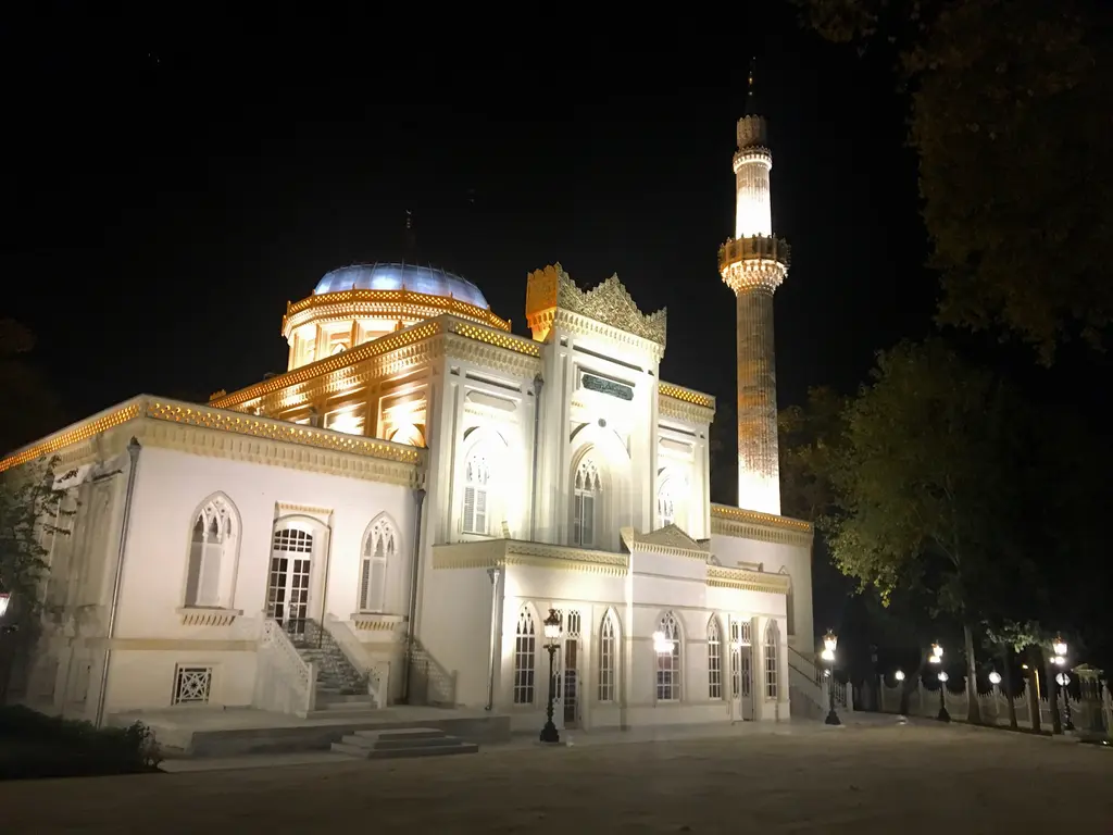Gece ışıklandırılmış Yıldız Hamidiye Camii; aydınlatılmış beyaz cephe ve kubbe kasnağı, parlayan minare ve karanlık ağaçlar.