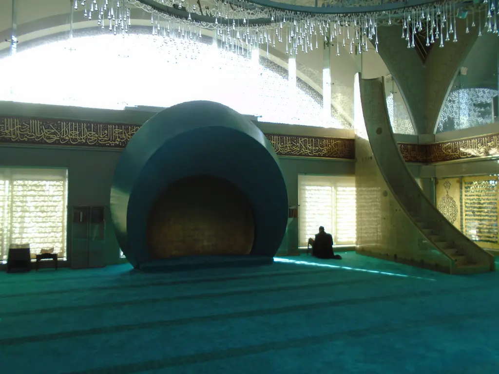 Şakirin Camii'nin harimi; turkuaz halı, kabuk biçimli mihrap, yanında minber ve arkada ışık alan cam paneller.