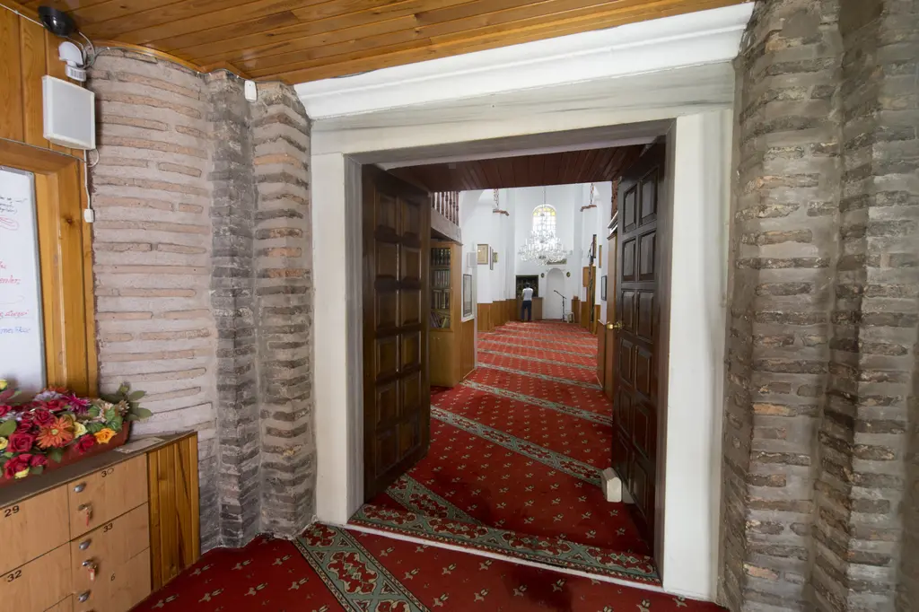 Bodrum Camii'nin narteksi; sıvası alınmış tuğla ve taş duvarlar, ahşap tavan, açık çift kanatlı koyu ahşap kapıdan harime bakış, solda ayakkabılık ve ilan panosu.