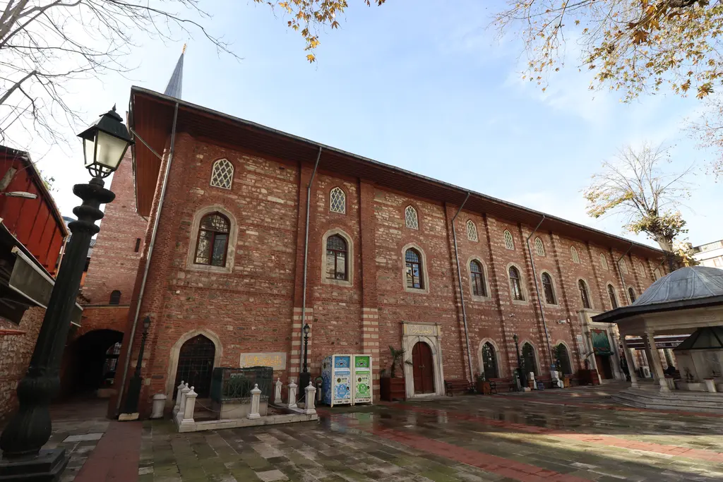 Galata'da Arap Camii; tuğla ve taş bantlı uzun cephe, sivri kemerli pencereler, avluda şadırvan, ağaçlar ve arkada kare minare.