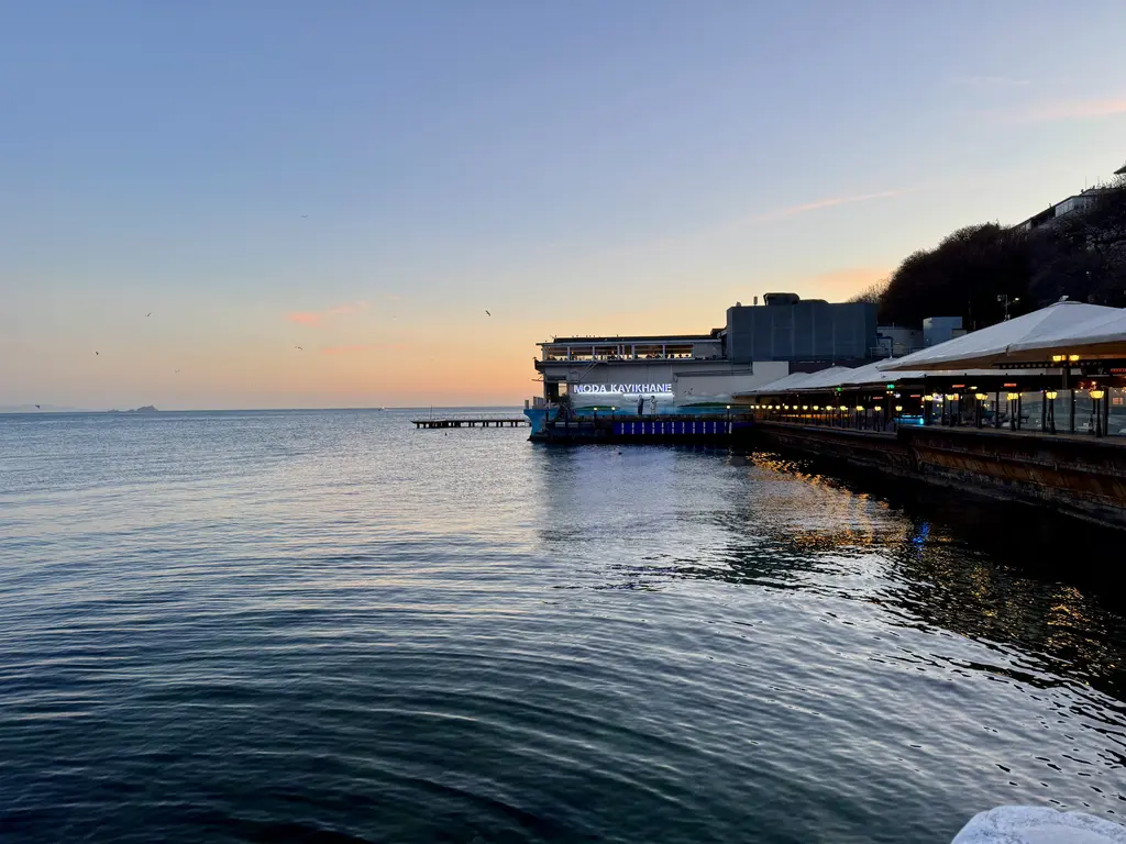Moda kıyısında akşam; sakin deniz, sağda ışıkları yanan kayıkhane ve restoran, ufukta pembe gökyüzü.