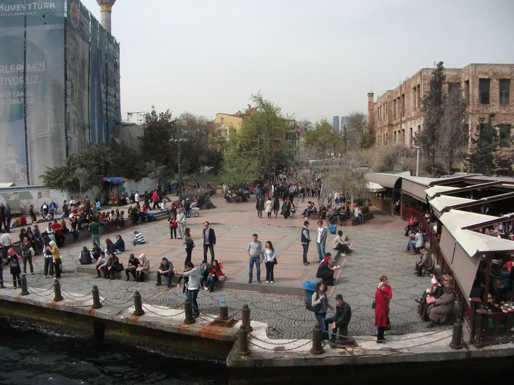 Ortaköy iskele meydanı ve arkada Esma Sultan Yalısı; taş döşeli kıyıda kalabalık, tenteli tezgâhlar ve ağaçlar.