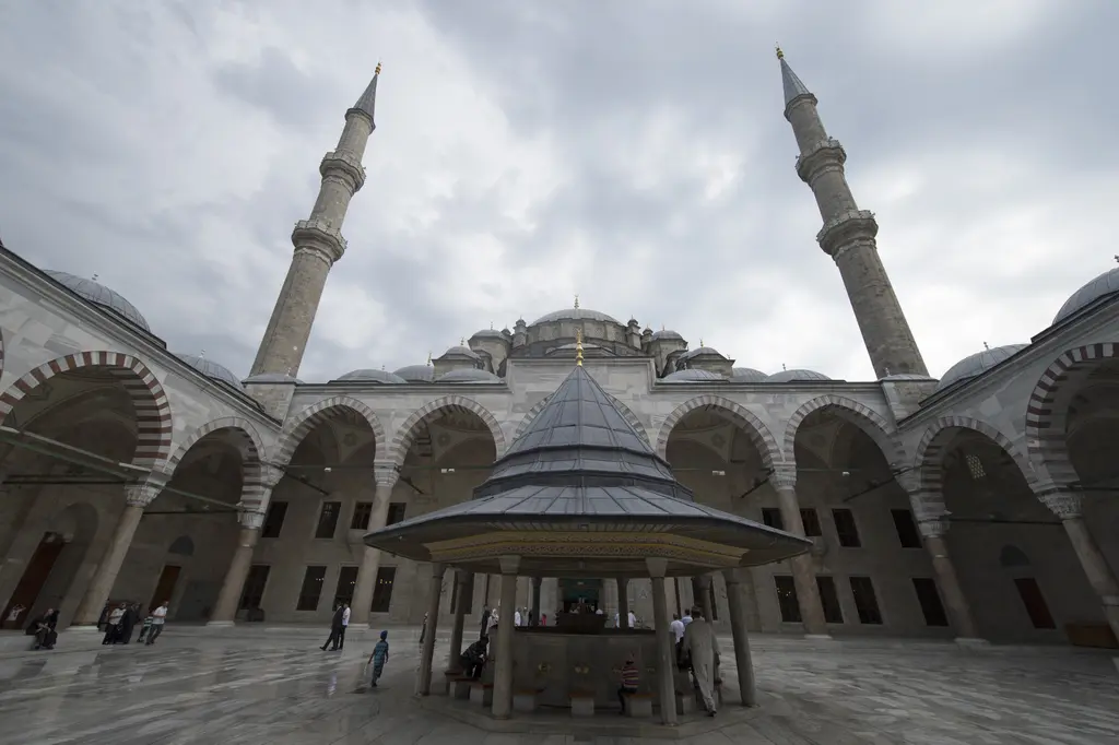 Fatih Camii'nin avlusu geniş açıyla; ağır gri bulutlar, iki yanda yükselen minareler, ortada konik kurşun çatılı şadırvan ve çevrede almaşık kemerli, mermer sütunlu revak.