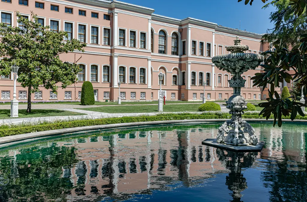 Dolmabahçe Sarayı bahçesinde havuz; ortada beyaz mermer kuğu figürleriyle süslü fıskiye, çevresinde yeşil su ve ağaçlar.
