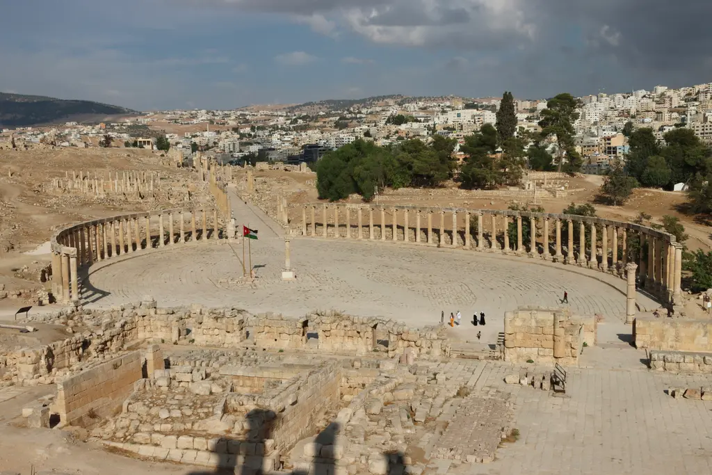 Jerash'ta yukarıdan görünen oval meydan; meydanı çevreleyen sütun dizisi, taş döşeli zemin, çevredeki antik kalıntılar ve arkada modern şehir ile yeşil tepeler.