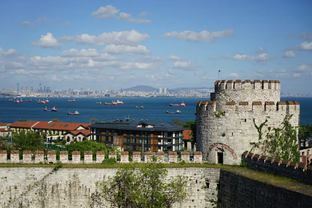 Yedikule Hisarı'nın kulesi ile mazgallı perde duvarı; solda Marmara Denizi'nde sıralanmış yük gemileri, uzakta şehir silueti ve üstte açık mavi gökyüzü.