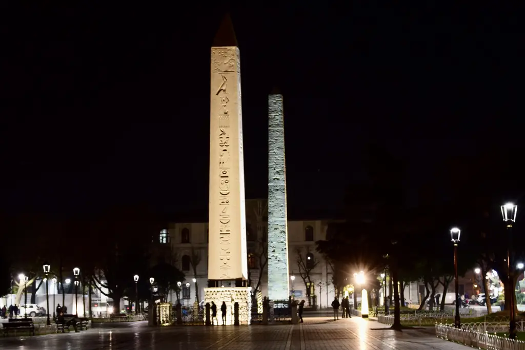 Gece aydınlatılmış Dikilitaş ve arkasındaki Örme Sütun; ıslak taş zemin, sokak lambaları ve meydanda yürüyen insanlar.
