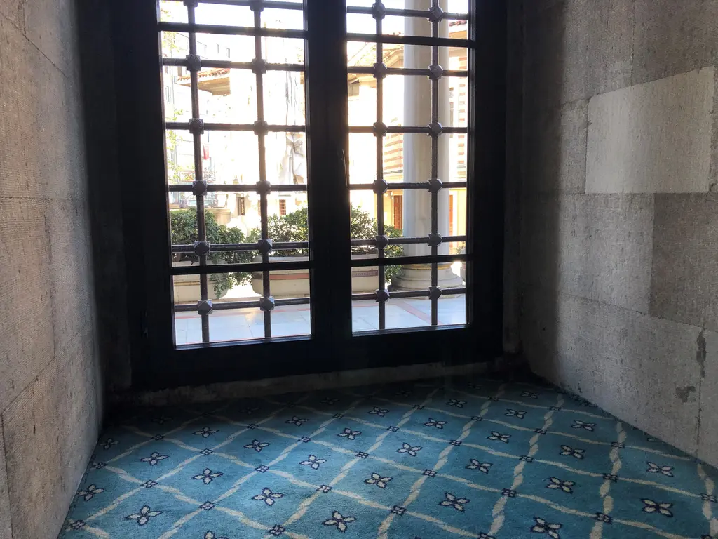 Şişli Camii'nin alt kat penceresi; kalın taş duvar içinde demir parmaklıklı pencere, dışarıda avlu ve mavi desenli halı.