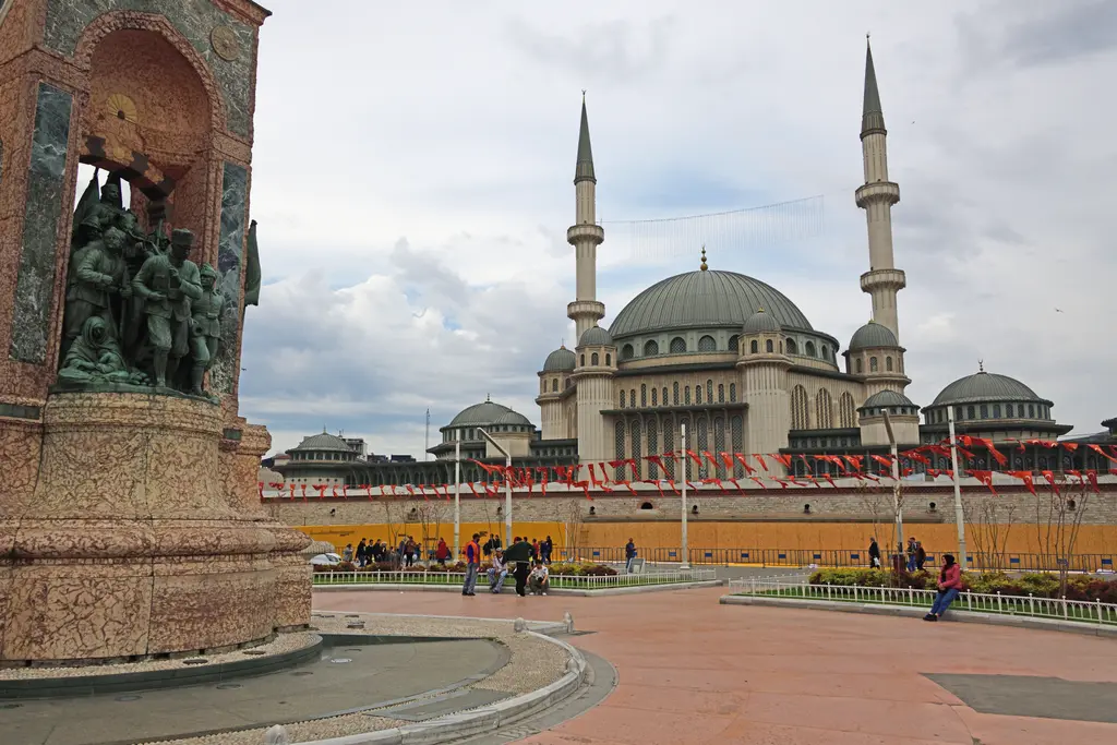 Taksim Meydanı'nda Cumhuriyet Anıtı ve arkasında Taksim Camii; solda bronz figürlü anıt, ortada taş zemin, sağda kubbeli cami.