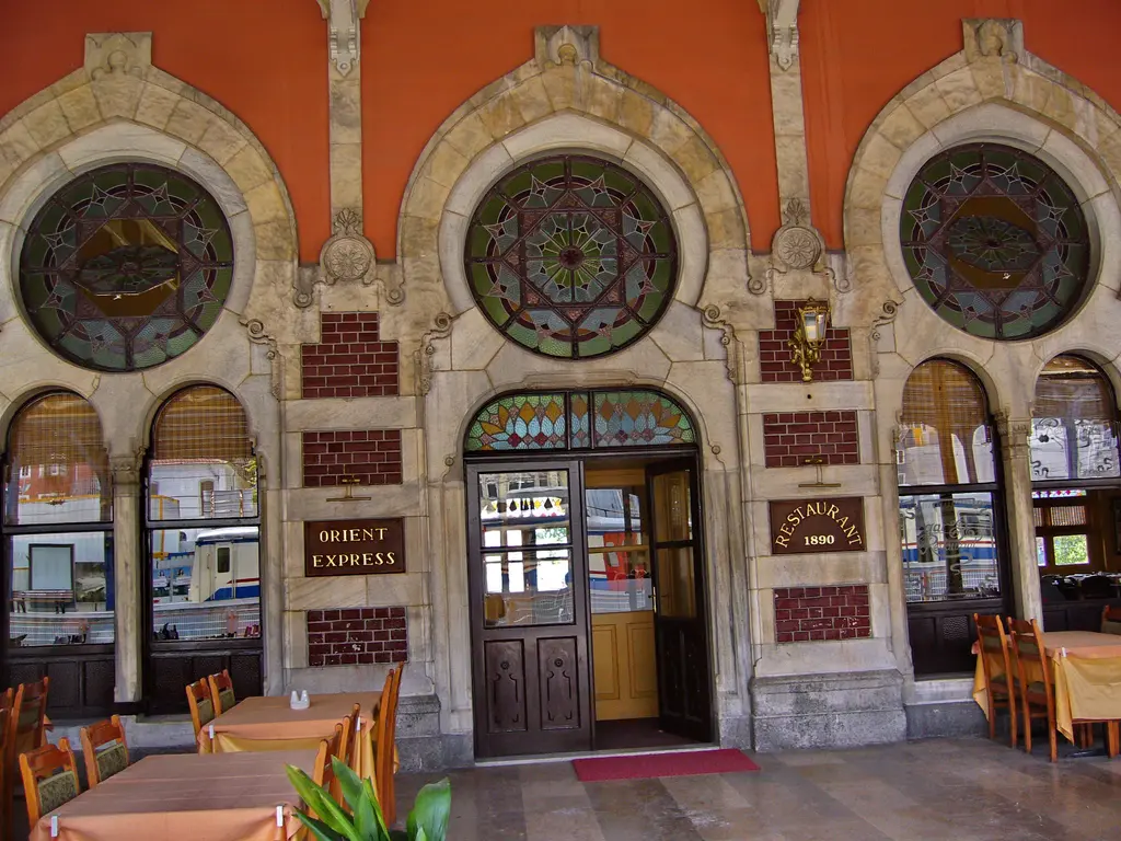 Sirkeci Garı'nda Orient Express lokantasının girişi; sol ayakta pirinç tabela, koyu ahşap kapı ve kırmızı paspas, üstte krem taş çerçeveli üç gül pencere, önde örtülü masalar.