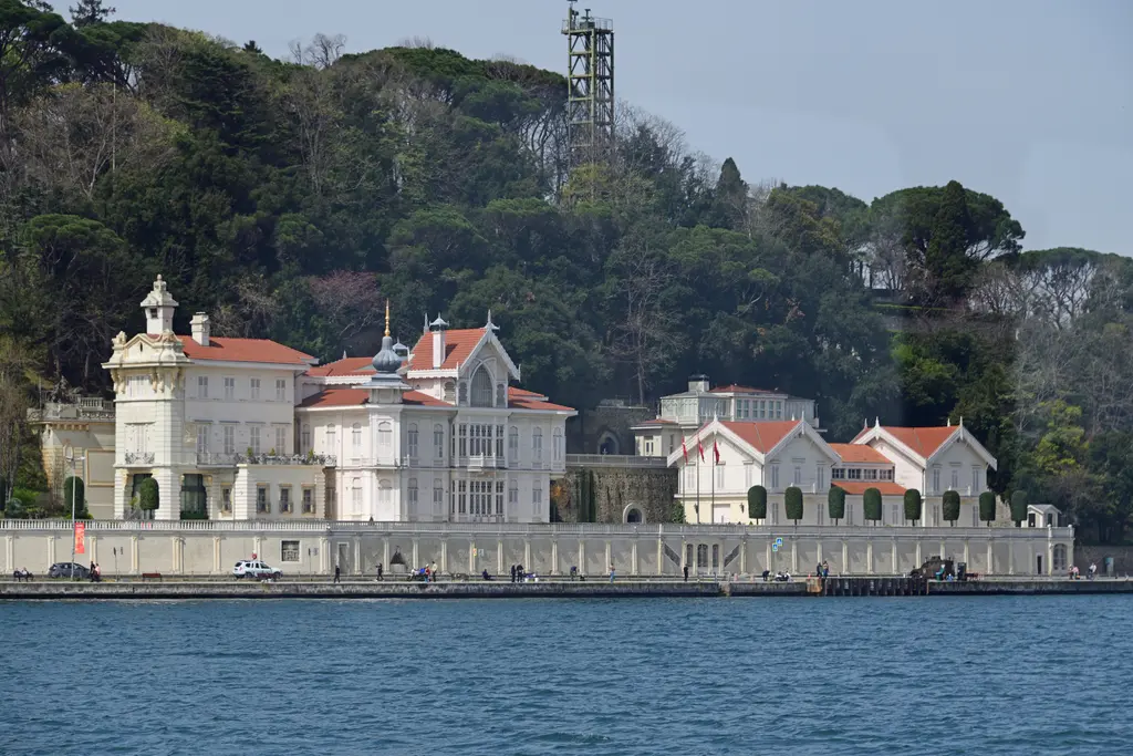 Huber Köşkü ve yan binaları; kıyıda beyaz ahşap yapılar, kiremit çatılar, arkada çam ağaçları ve kule.