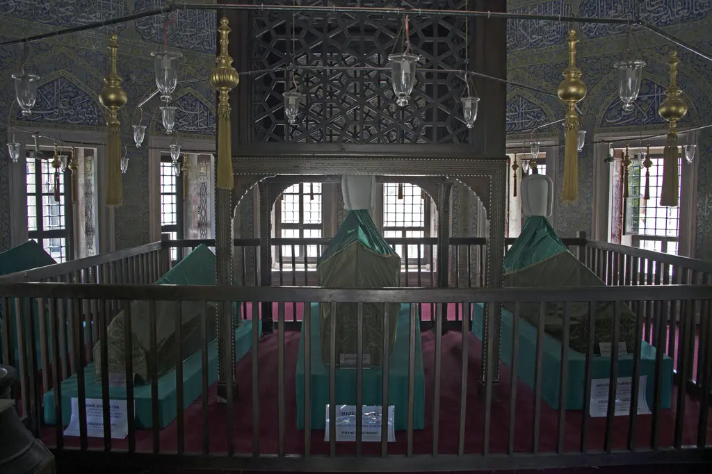 Şehzade Mehmed Türbesi'nin içi; parmaklığın ardında yeşil örtülü sandukalar, yukarıda kafesli ahşap örtü, asılı pirinç kandiller ve loş ışık.