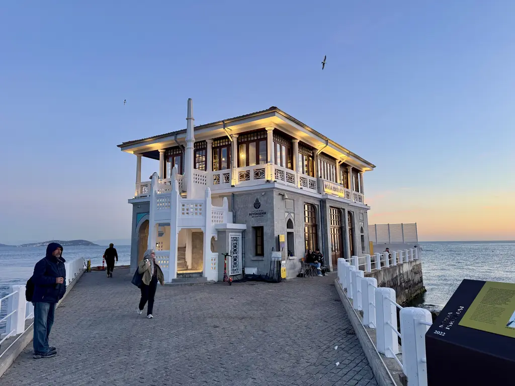Moda İskelesi; ışıkları yanan iki katlı bina, beyaz korkuluklu taş yol, yürüyen insanlar ve alacakaranlık gökyüzü.