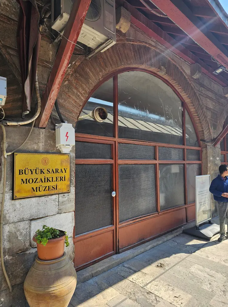 Arasta Çarşısı'nda kemerli bir kapı; üstünde sarı zemin üzerine "Büyük Saray Mozaikleri Müzesi" yazan levha ve önünde bir küp.