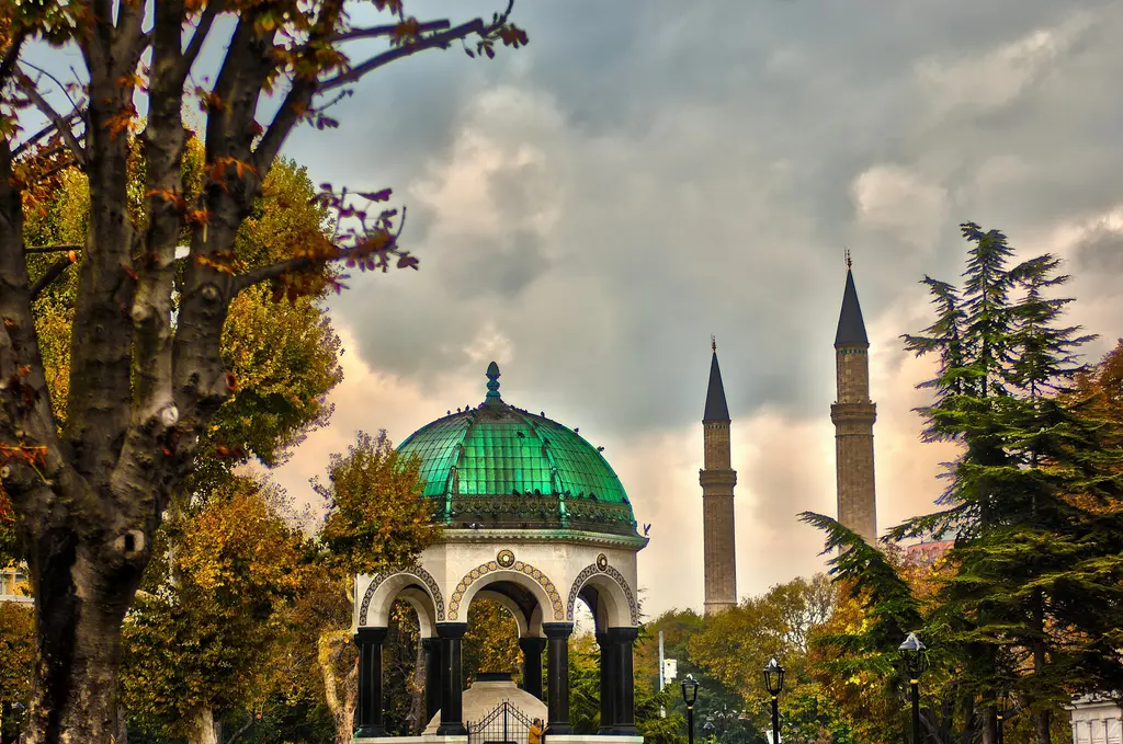 Sonbaharda Alman Çeşmesi; yeşil kubbe, sarı yapraklı ağaçlar, arkada Sultanahmet Camii'nin minareleri ve bulutlu akşam gökyüzü.