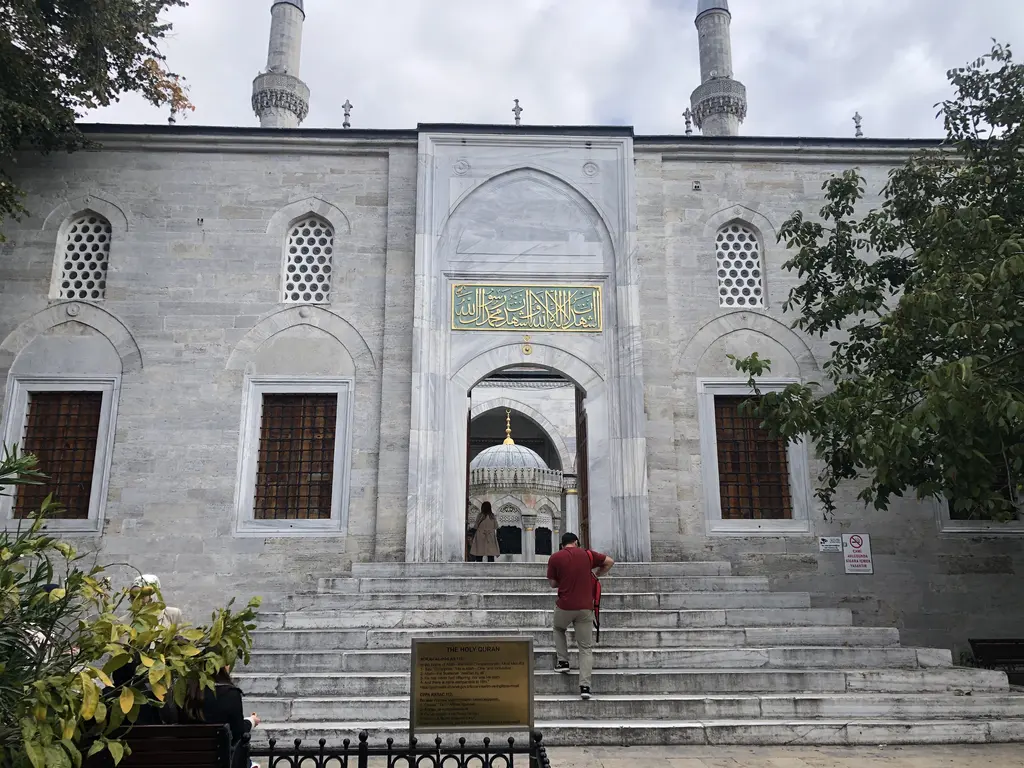 Yeni Valide Camii'nin avlu girişi; mermer cephe, yaldızlı yazı levhası, geniş merdiven ve çıkan bir ziyaretçi.