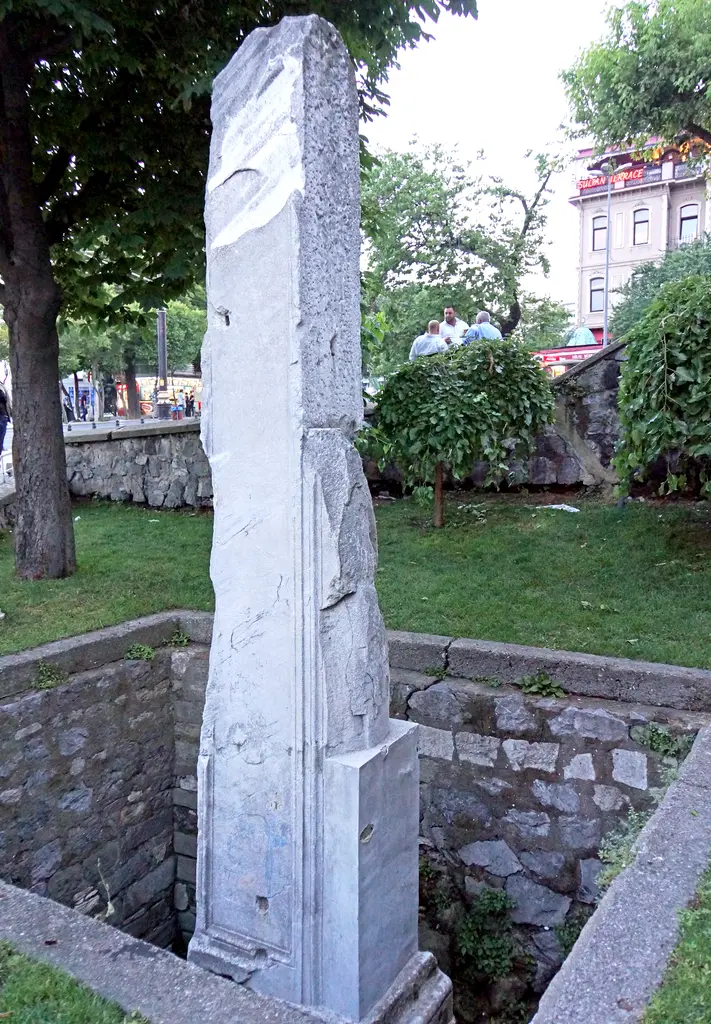 Milyon Taşı; taş duvarlı çukurun içinden yükselen açık renk mermer ayak, çevresinde çimen, arkada ağaçlar ve kalabalık.