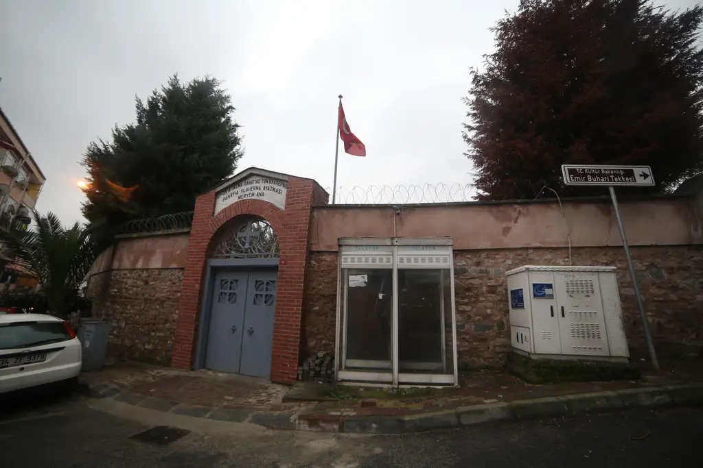 Sokaktan bahçe duvarı; kırmızı tuğlalı kemerli gri kapı, yanında elektrik dolapları, bayrak direği ve duvarın ardında yüksek ağaçlar.