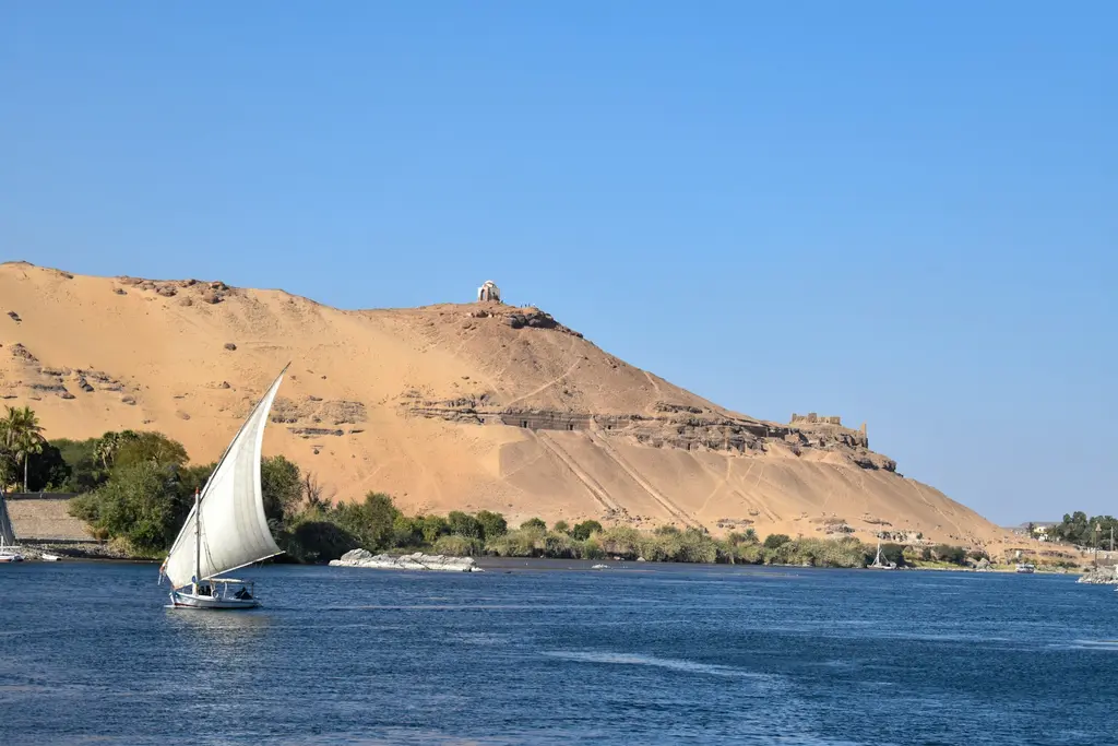 Mısır'da Asvan yakınında Nil Nehri; yelkenini açmış geleneksel bir felucca teknesi ve karşı kıyıdaki sarı kumul tepe, berrak öğle ışığı.