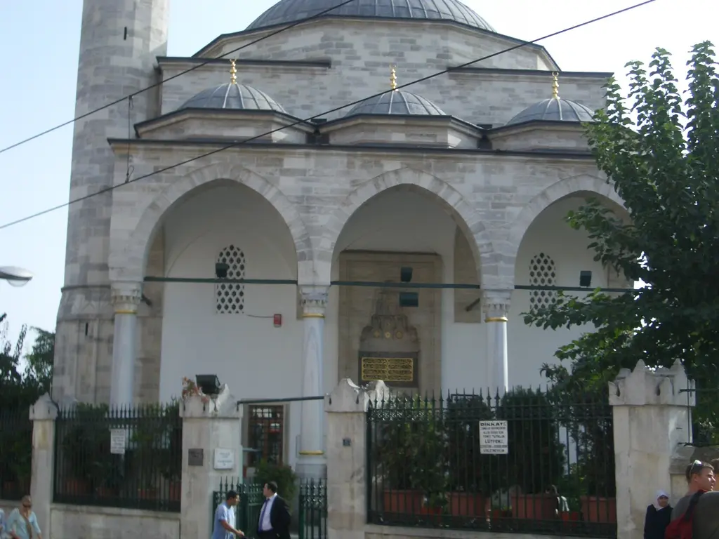 Firuz Ağa Camii'nin ön cephesi; kemerli revak, küçük kubbeler, ana kubbe, önde demir parmaklıklı avlu duvarı ve bitkiler.