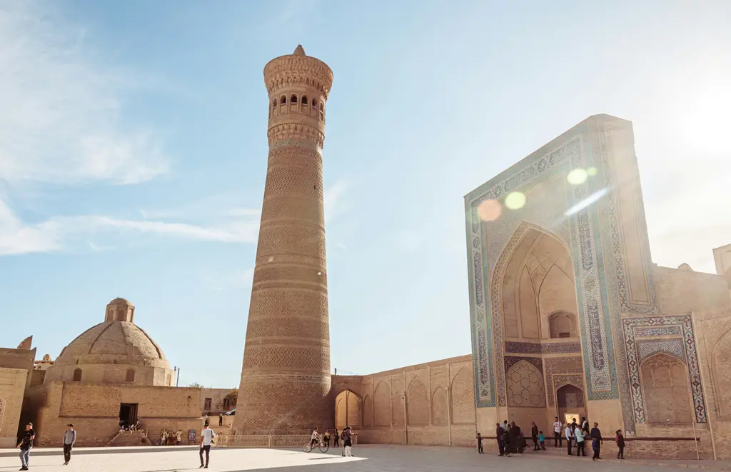 Buhara'da Poi Kalyan meydanında yükselen tuğla Kalyan Minaresi; yanında mavi kubbeli cami ve medrese portalleri.