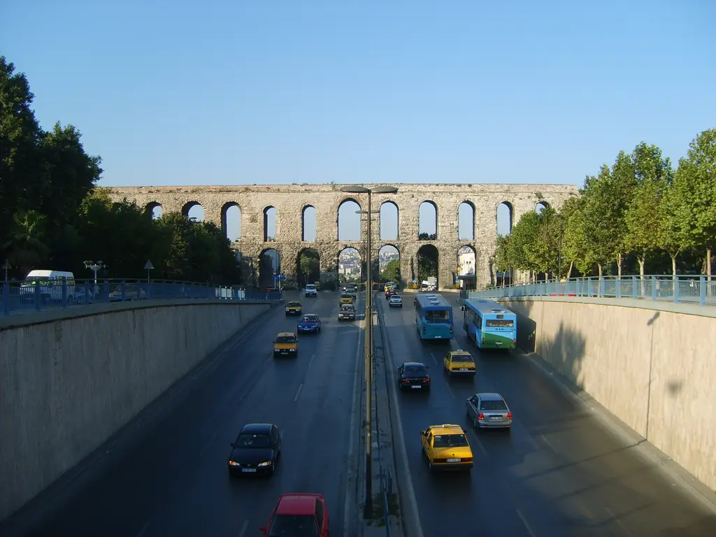 Bozdoğan Kemeri'ne bulvar boyunca uzaktan bakış; önde beton istinat duvarları arasında inen yol ve araçlar, uzakta kemer yolu enlemesine kesiyor, iki yanda ağaç sıraları.