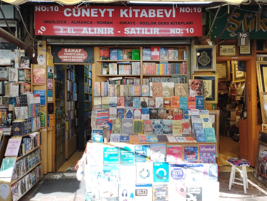 Sahaflar Çarşısı'nda kırmızı zeminli bir kitabevi tabelası; İngilizce, Almanca, roman, sözlük ve ders kitabı sıralaması, altında renkli kitap standları, sağ duvarda hat levhaları.
