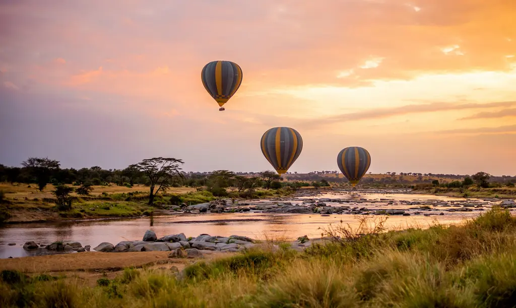 Serengeti'de gün doğumunda bir nehrin üzerinde süzülen üç sıcak hava balonu; pastel turuncu-mor gökyüzü, kayalık nehir yatağı ve akasyalar.