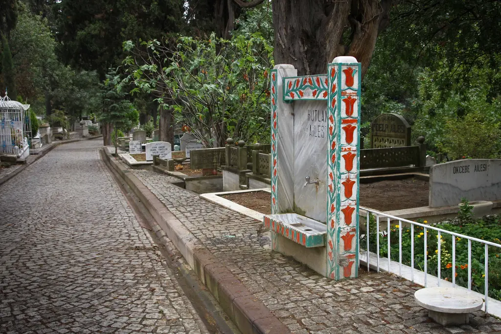 Karacaahmet'te boyalı çeşme; renkli desenli taş gövde, beyaz parmaklık, yanında parke taşı yol ve ağaçlar.