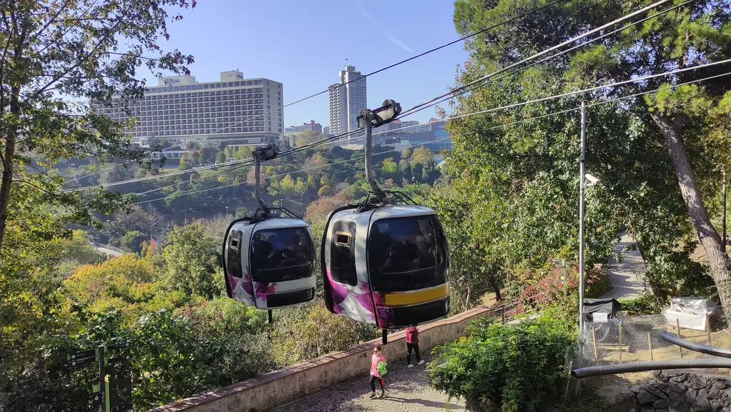 Maçka Demokrasi Parkı'nın üstünde iki beyaz mor teleferik kabini; altta ağaçlı vadi ve taş yol, arkada sonbahar ağaçları ardında Hilton.