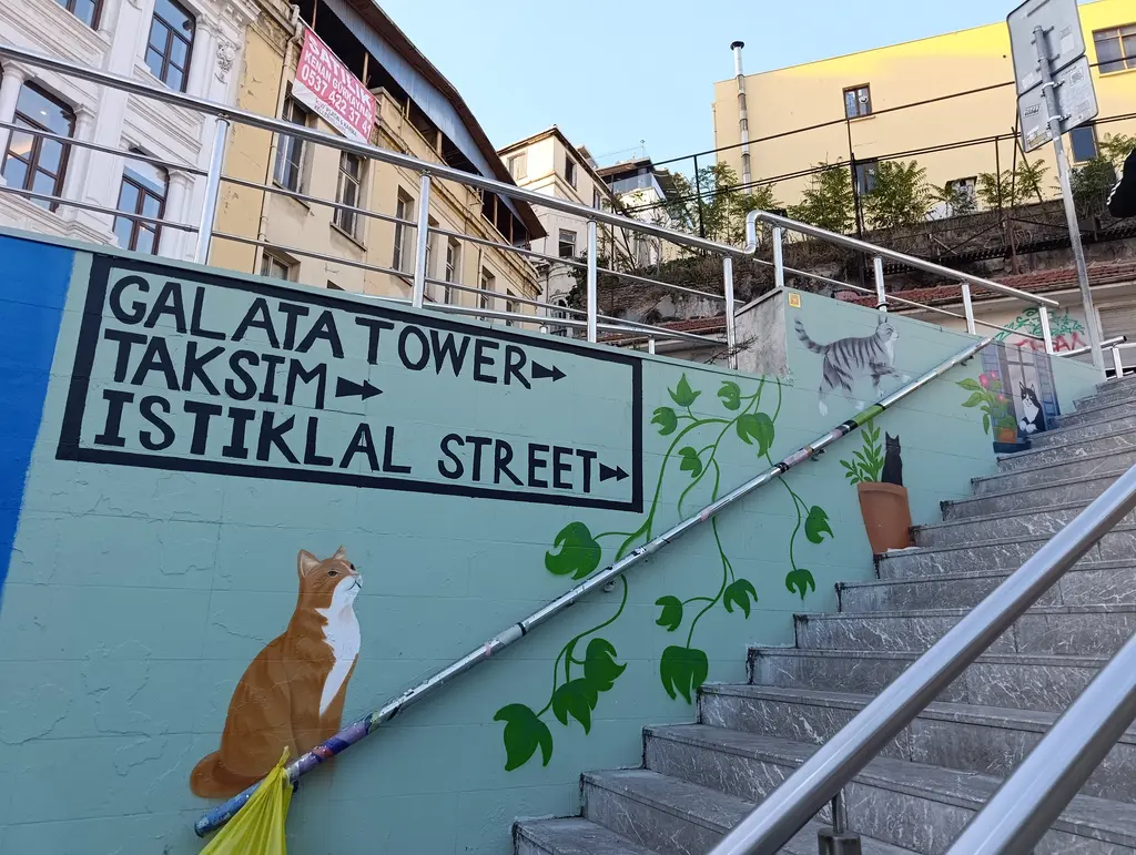 Karaköy alt geçidinin girişi; duvarda kedi resmi, Galata Tower, Taksim ve Istiklal Street yazılı tabela ve merdiven.