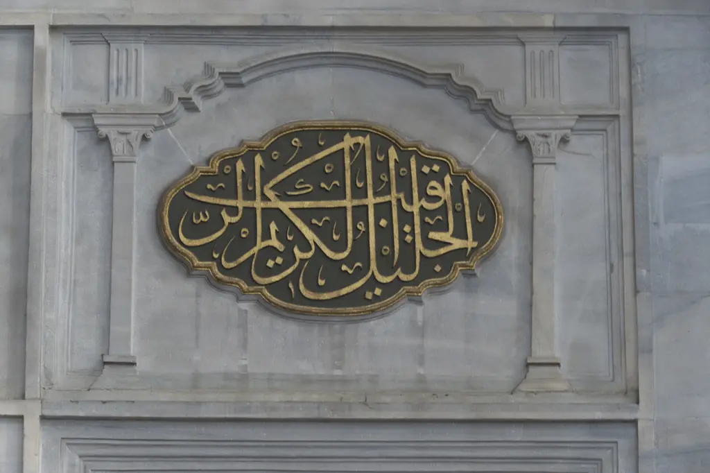 Nuruosmaniye Camii'nde beyaz mermer duvara oyulmuş çerçeve içinde oval hat levhası; yaldızlı harfler, çevresinde kabartma silme ve eşit dağılan ışık.