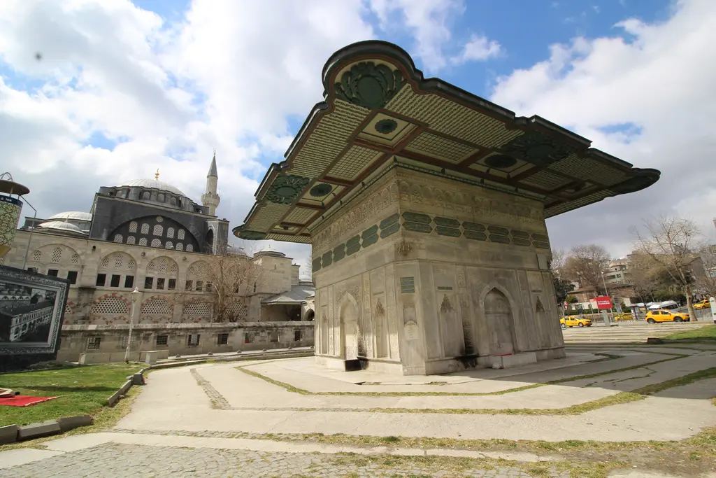 Tophane Meydanı; ortada geniş saçaklı çeşme, sağda Kılıç Ali Paşa Camii'nin kubbeleri ve revakı, önde taş döşeli yollar ve çimen.