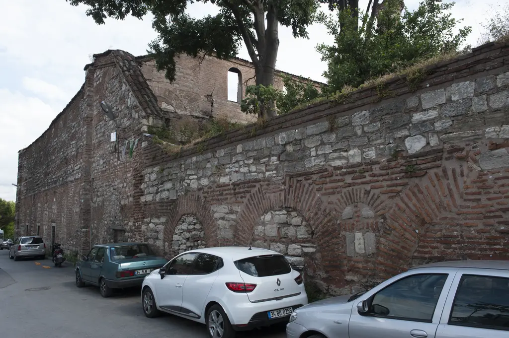 İmrahor İlyas Bey Camii'nin sokak cephesi; boydan boya uzanan almaşık örgülü duvar, üstünde yıkık bina kütlesi ve ağaçlar, önde park etmiş arabalar.