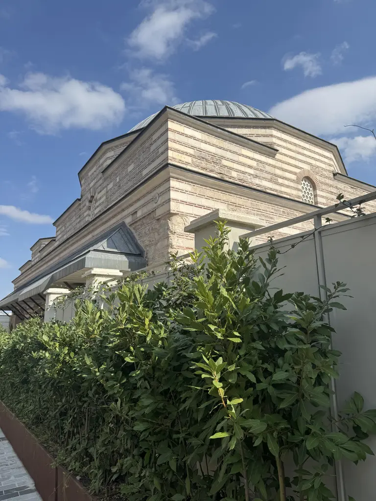 Çinili Hamam'ın kubbesi aşağıdan; kurşun kaplama, altında kesme taş cephe, önde yeşil çalı sırası ile cam korkuluk, bulutlu mavi gök.