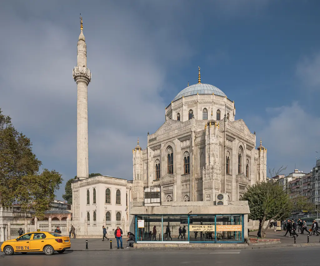 Pertevniyal Valide Sultan Camii ve önündeki beyaz türbe; ince minare, kubbe, cadde kenarında sarı taksi ve otobüs durağı.