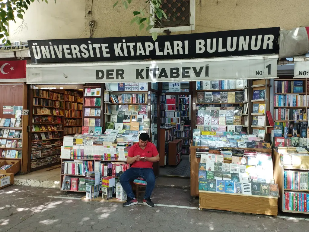 Sahaflar Çarşısı'nda "Üniversite kitapları bulunur" yazılı bir dükkân cephesi; solda küçük Türk bayrağı, önünde kutunun üstüne oturmuş telefonuna bakan genç ve üç sıra kitap standı.
