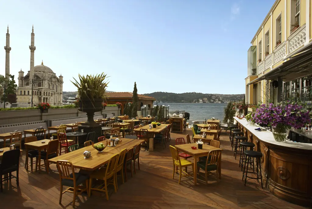 Ortaköy'de kıyıdaki bir kafe terası; ahşap masalar, çiçekli saksılar, arkada cami ve minareleri ile Boğaz.